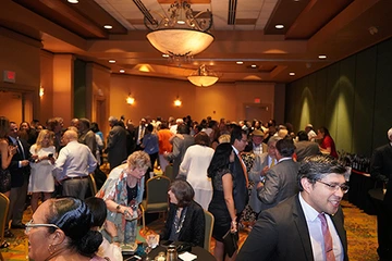 A large crowd of guests socializing and mingling at a formal indoor reception in a brightly lit, carpeted banquet hall with warm overhead lighting.